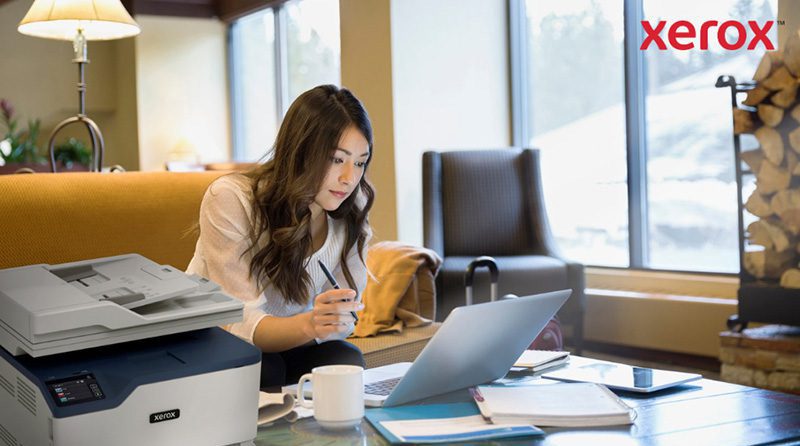 Una donna lavora al computer portatile in un comodo ufficio o in una sala relax, con una stampante compatta Xerox visibile in primo piano, a indicare la perfetta integrazione della stampa con i flussi di lavoro digitali.