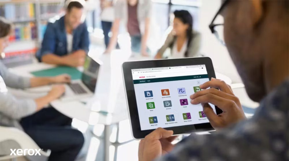 Una persona tiene in mano un tablet che mostra l'interfaccia di Xerox Workflow Central con varie icone di app, mentre altre persone lavorano sullo sfondo sfocato, evidenziando l'accesso mobile alle soluzioni di flusso di lavoro.