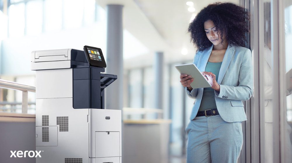Una persona è in piedi accanto a una stampante multifunzione Xerox VersaLink B605, osserva e interagisce con un dispositivo tablet, illustrando come ConnectKey consente l'interazione mobile con la stampante.
