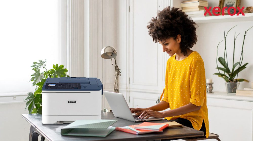 Xerox® DocuShare® Go: produttività per il lavoro ibrido Una persona con i capelli ricci sorride mentre scrive su un computer portatile seduto alla scrivania, con una stampante compatta Xerox C310 posizionata accanto al computer portatile, a dimostrazione di una moderna configurazione di ufficio domestico per la produttività.