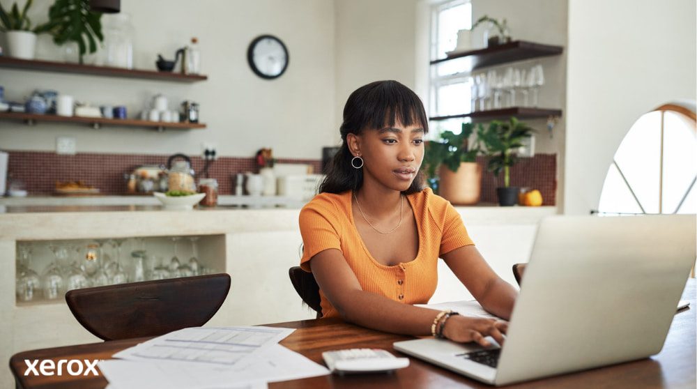 Xerox® DocuShare® Go: potenziare il lavoro da remoto Una persona è seduta al tavolo della cucina, concentrata sul lavoro con un computer portatile, con documenti e una calcolatrice a portata di mano, a dimostrazione della flessibilità del lavoro da casa con una gestione efficiente dei documenti.