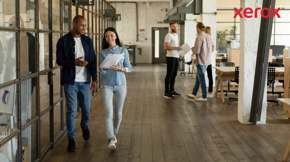 Due colleghi camminano lungo un corridoio in un moderno ufficio open space, tenendo in mano dei documenti e sorridendo, con altri colleghi visibili sullo sfondo, a rappresentare un ambiente di lavoro dinamico e collaborativo.