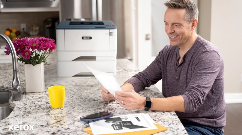 Una persona sorride mentre esamina un documento seduto al bancone della cucina, con una stampante compatta Xerox e una tazza gialla brillante nelle vicinanze, a suggerire una stampa e una gestione dei documenti senza problemi in un ambiente domestico o in un piccolo ufficio.