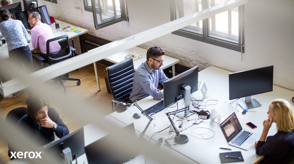 Una vista dall'alto di un ufficio open space con diverse persone che lavorano alle loro scrivanie, utilizzando i computer. La scena trasmette un ambiente di lavoro produttivo e collaborativo.