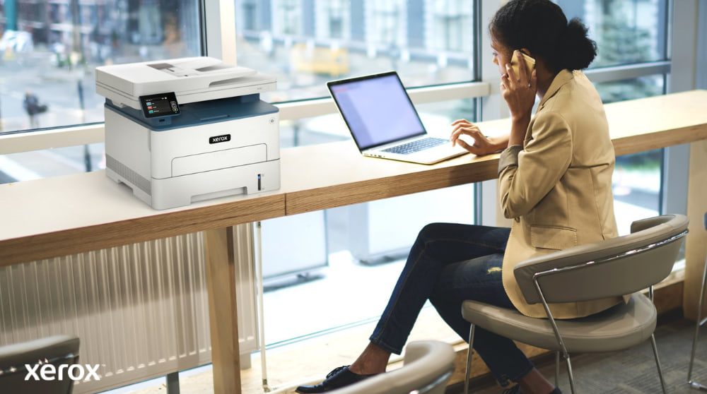 Una persona è seduta a un tavolo alto vicino a una grande finestra, mentre parla al telefono e lavora al computer portatile. Una stampante multifunzione Xerox B235DNI è comodamente posizionata sul tavolo accanto, a suggerire la facilità d'uso in un ambiente di lavoro moderno.