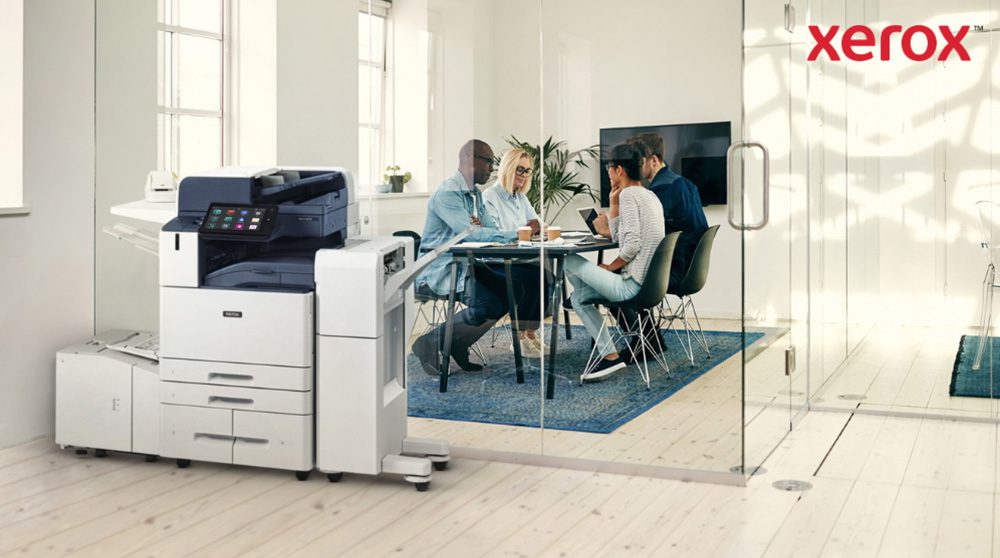 Una moderna stampante multifunzione Xerox AltaLink B8245 è in primo piano in un ufficio luminoso. Sullo sfondo, un gruppo di persone è impegnato in una discussione collaborativa attorno a un tavolo.