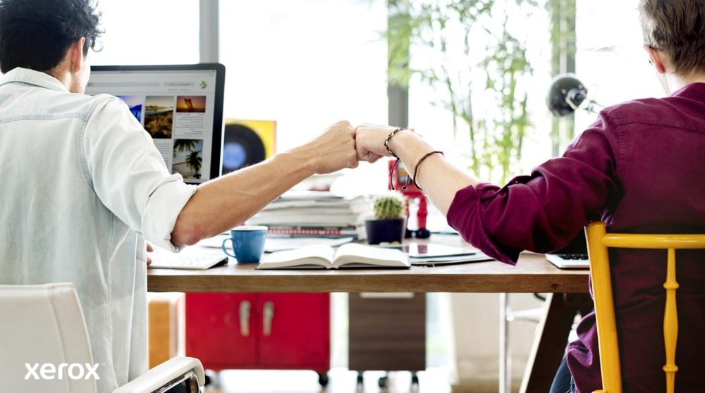 Due persone sedute a una scrivania, con le spalle rivolte alla telecamera, si salutano con un pugno, simbolo di lavoro di squadra e collaborazione di successo. Una di loro guarda lo schermo di un computer.