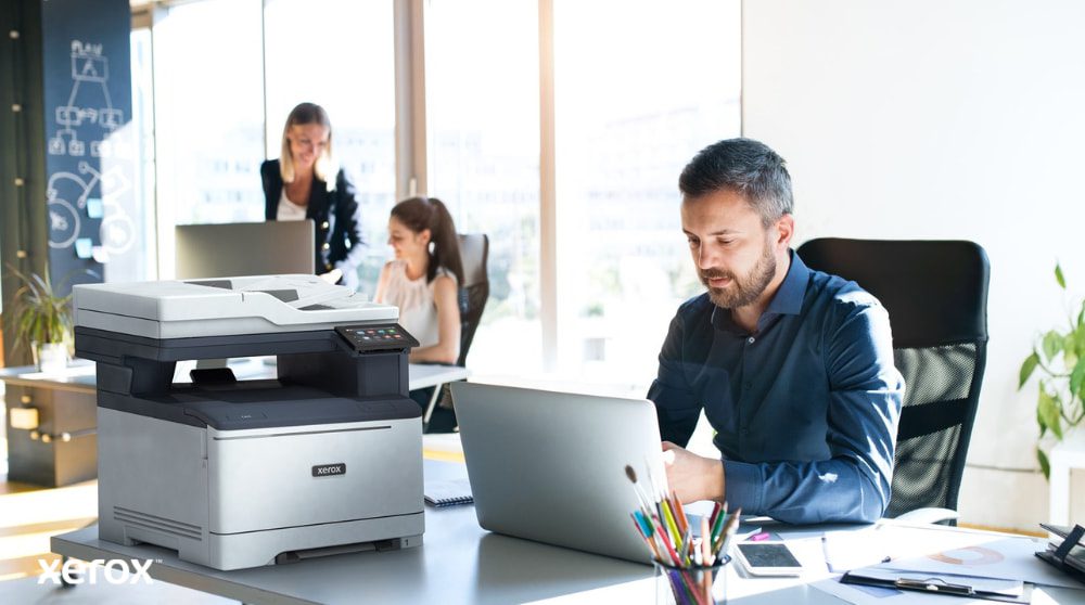 Una persona è seduta a una scrivania e guarda un computer portatile con accanto una stampante multifunzione Xerox C325. Sullo sfondo, altre due persone lavorano alle loro scrivanie in un ambiente luminoso.