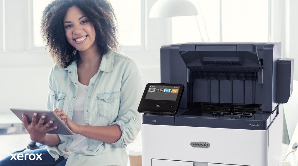 Una persona sorridente è seduta accanto a una stampante bianca Xerox VersaLink B610, tiene in mano un tablet e guarda la telecamera, evidenziando la natura intuitiva delle soluzioni Xerox.