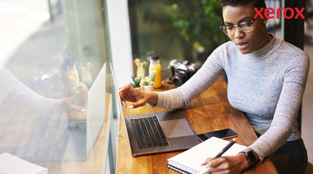 Una persona che indossa gli occhiali lavora a un tavolo di legno, guardando lo schermo di un laptop e scrivendo su un quaderno, con uno smartphone nelle vicinanze, suggerendo un ambiente di lavoro produttivo