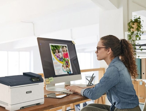 Una donna seduta alla scrivania, concentrata sul lavoro, con una stampante Xerox® C320 a colori